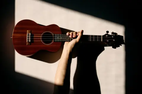 Dłoń trzymająca drewniane ukulele na jasnym tle z geometrycznymi cieniami.